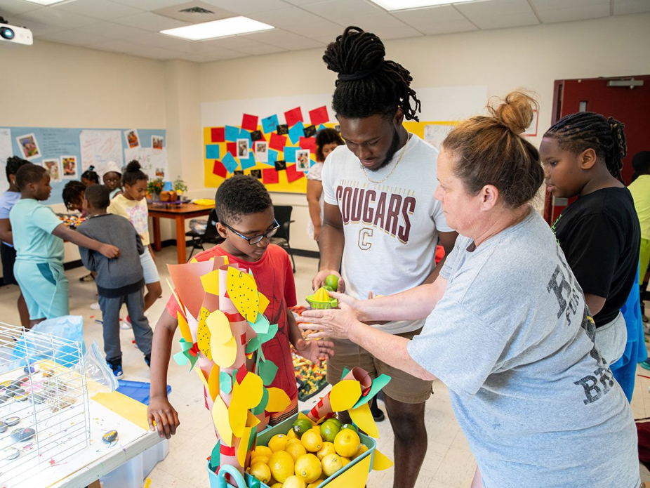 Inside an elementary school classroom, a male college student assists a female elementary school teacher in supporting her students' construction paper art projects.
