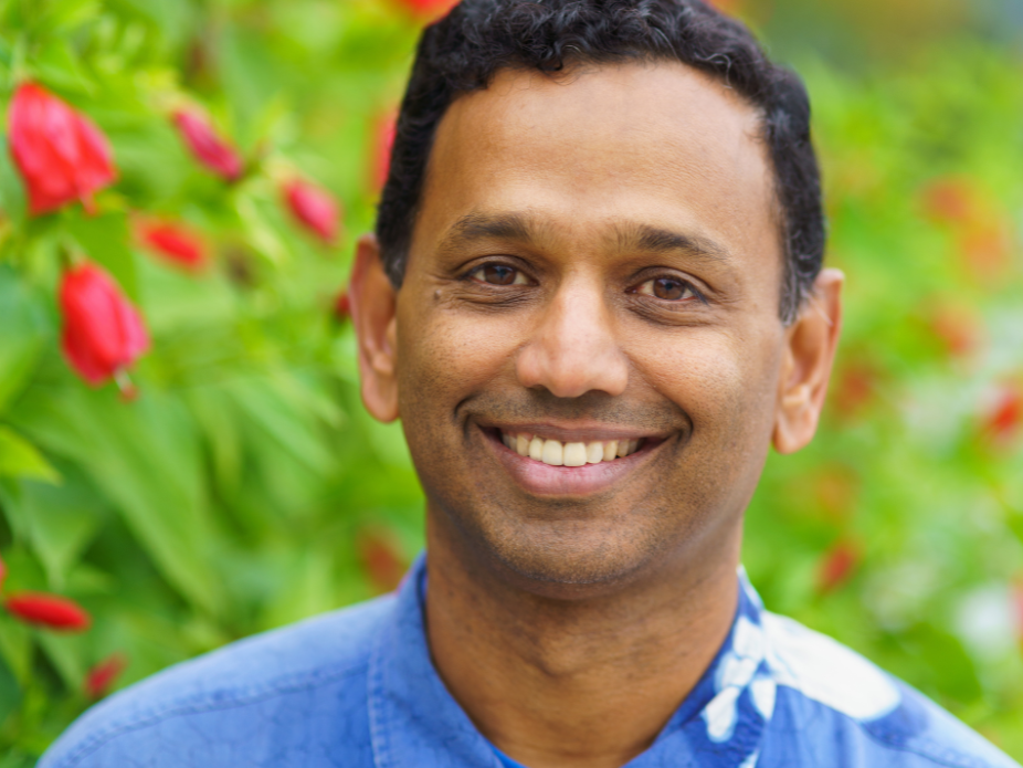 A smiling man wearing a blue Hawaiian shirt stands foregrounded in front of foliage and red flowers.