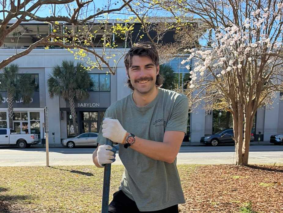 Person wearing a gray T-shirt and white gloves is holding a shovel in an outdoor area with trees and mulch. A row of storefronts and parked cars is visible in the background under a clear blue sky.