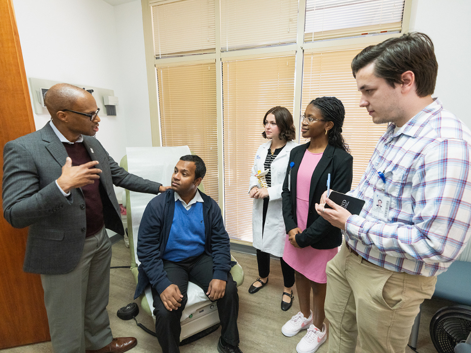 A group of five people is gathered in a medical office. On the left, a person in a suit and glasses is speaking to the group. Seated in front of them is an individual in a blue sweater and collared shirt, looking up attentively. Standing nearby are three others: one in a white lab coat holding papers, another in a pink dress and black blazer with a pen and notebook, and a third in casual attire with an ID badge. Large windows with partially closed blinds are visible in the background.