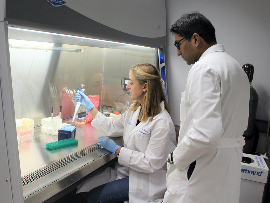 Two individuals in white lab coats and blue gloves working in a laboratory. One person is seated under a fume hood using a pipette to transfer liquid into a container while the other person stands nearby observing.