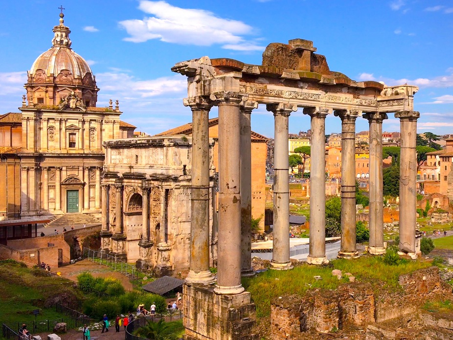 Ancient stone columns stand among ruins in a historic city site, with a domed church and additional buildings visible in the background under a blue sky.