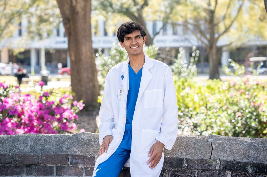 Ronit Pathak, a triple-major in international studies, chemistry, and biochemistry, sits against a brick wall while wearing a medical coat.