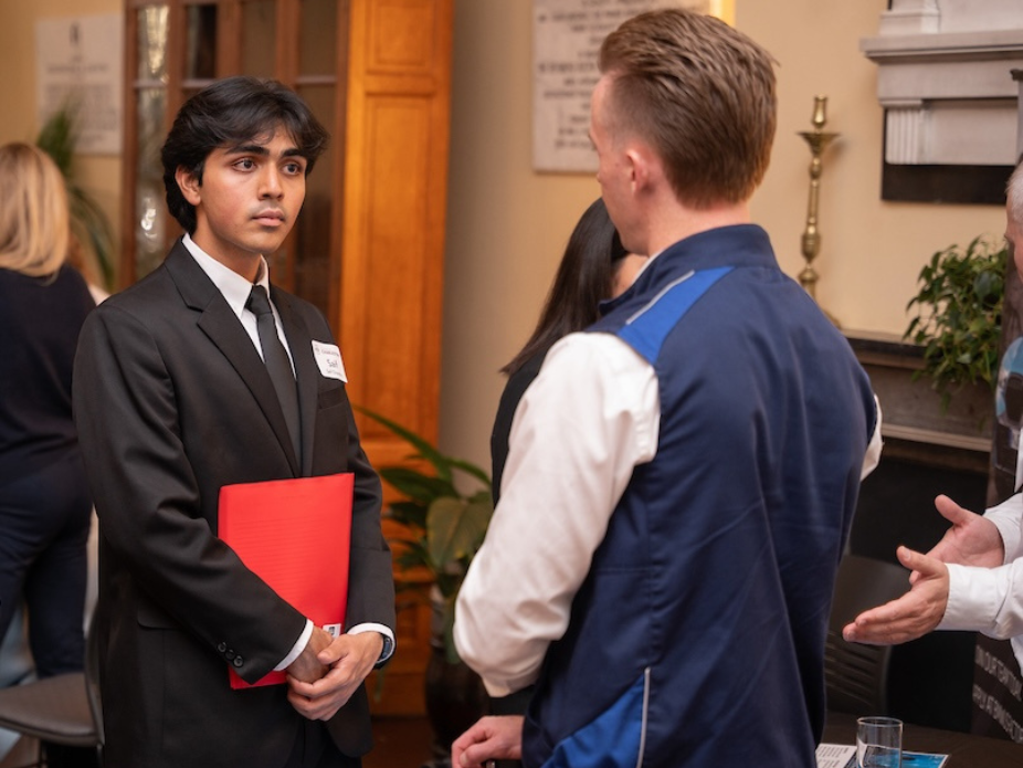 A young person in a black suit and tie holding a red folder is conversing with another individual in a blue jacket at an indoor event. The background includes other attendees, wooden furniture, potted plants, and framed documents on the wall.