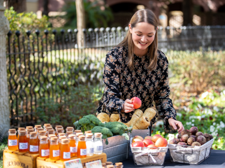  A person stands behind an outdoor market table arranging fresh produce, including apples, root vegetables, and broccoli. Bottled drinks are neatly lined up on one side of the table, with a garden fence and greenery behind.