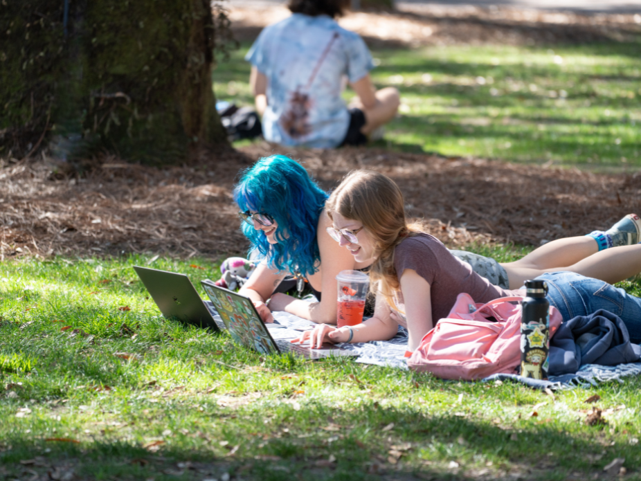  Two people lie on a blanket on a grassy campus lawn, working side by side on open laptops. A backpack, water bottle, and iced drink rest nearby, with trees and other people relaxing in the background.