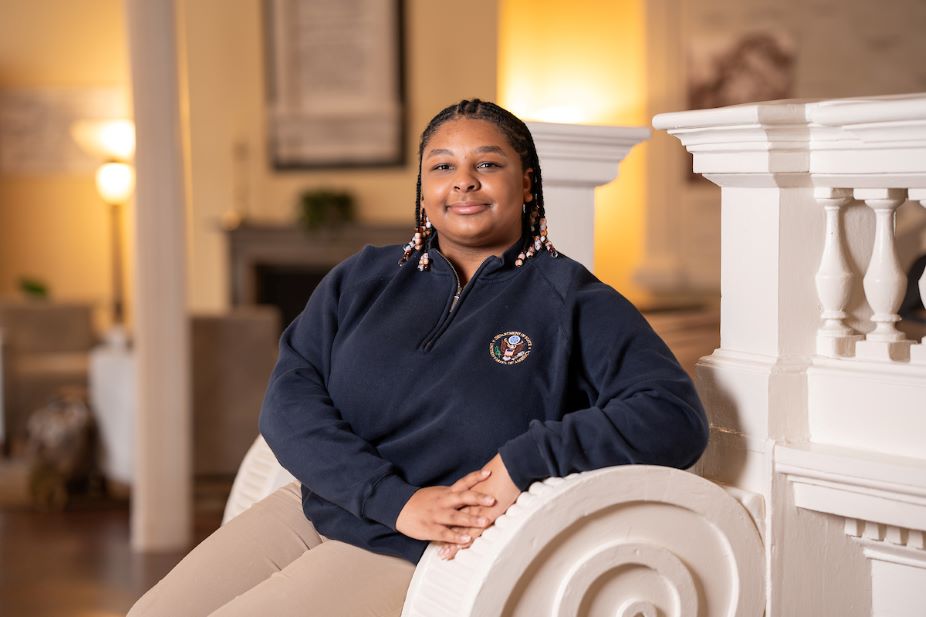 Syd Jackson, a double-major in international studies and political science, leans against a post while wearing a state department pullover.