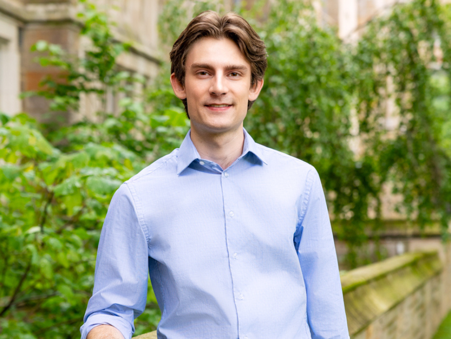A young man in a blue button-down shirt poses in front of a building.