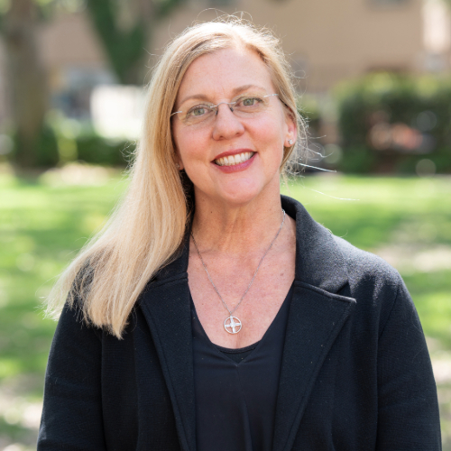 A woman with glasses and black clothes smiling outside on a sunny day