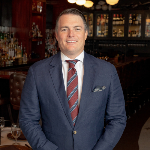 Professional portrait of a man in a suit standing in a bar setting with a well-stocked liquor display in the background.