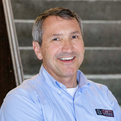 Smiling man wearing a blue checkered shirt stands in front of a staircase, conveying professionalism and approachability.
