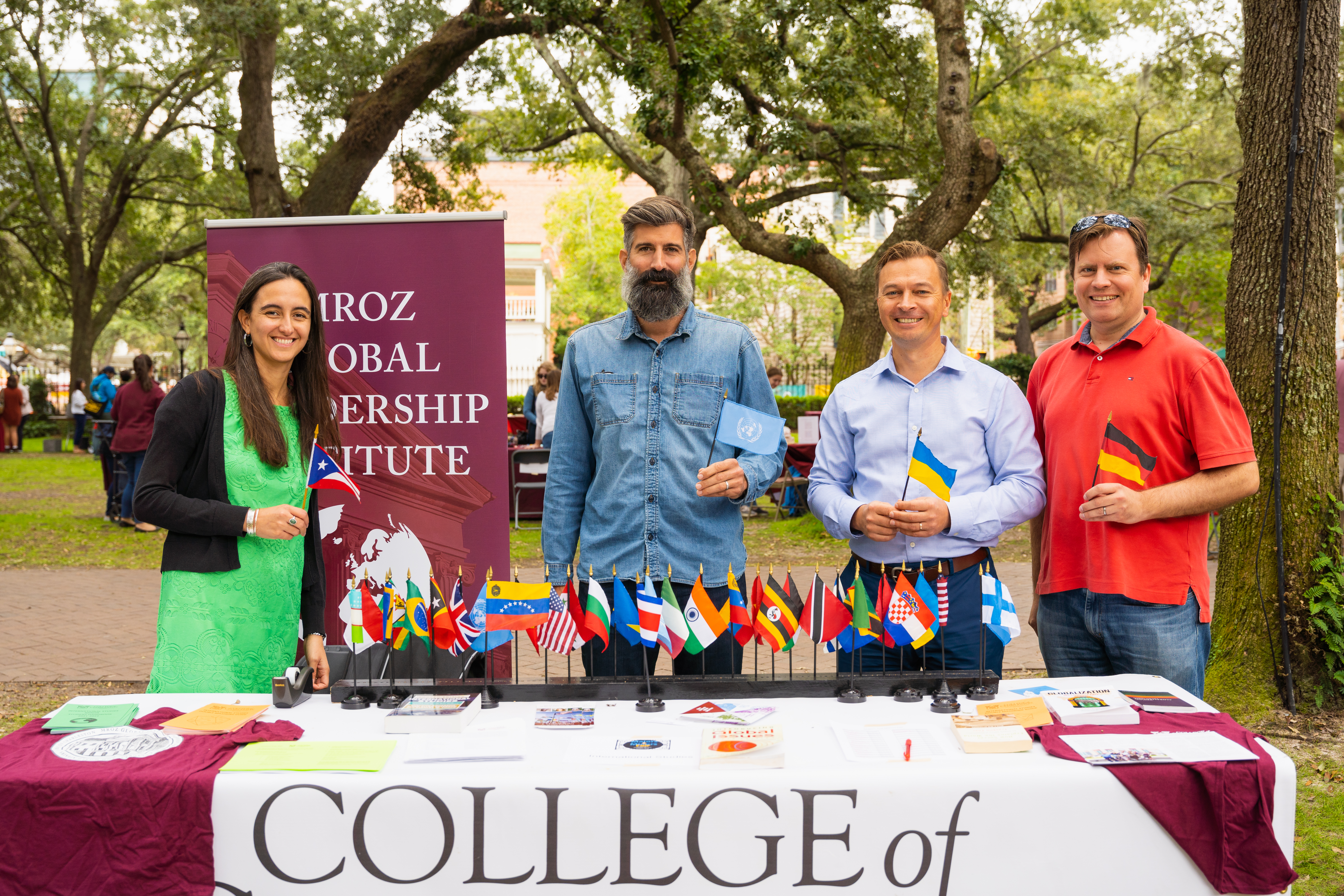 One female and three male professors (Drs. Bea Maldonado, Blake Scott, Max Kovalov, and Malte Pehl, respectively) standing behind a table with a rack of mini flags of countries from around the world.