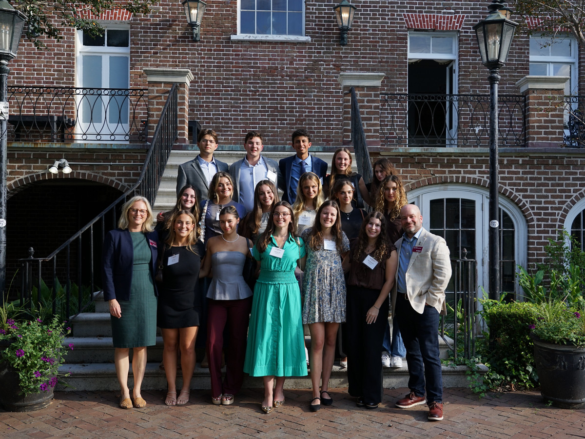 16 Perlmutter Fellows flanked by their 2 faculty advisors standing on the stairs of a historic brick house and smiling.
