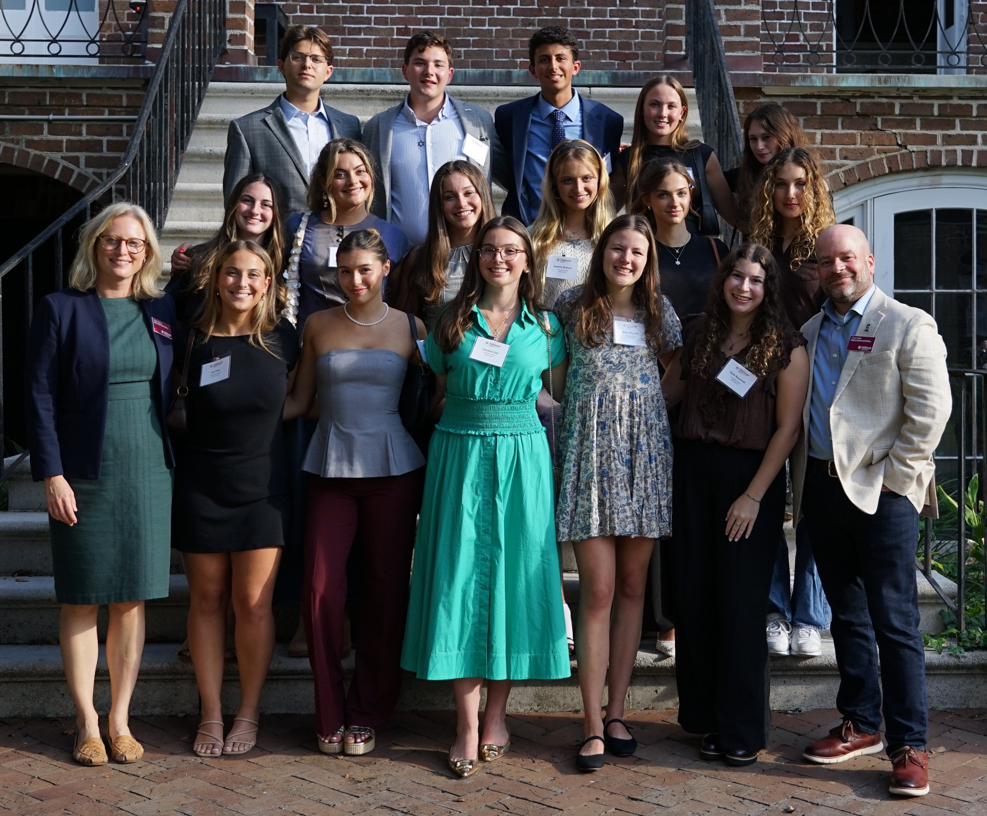 16 Perlmutter Fellows flanked by their 2 faculty advisors standing on the stairs of a historic brick house and smiling.