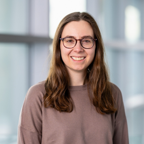 Headshot of a person with glasses and long brown hair, smiling against a soft office background.