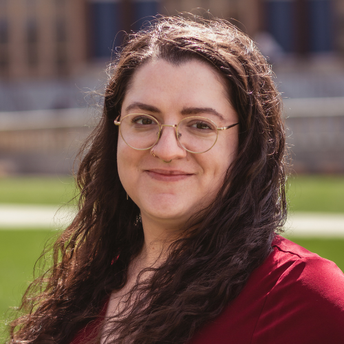 Portrait headshot of a person with long wavy dark hair and round glasses, wearing a red top, standing outdoors on a sunny day with a blurred campus lawn and building in the background.
