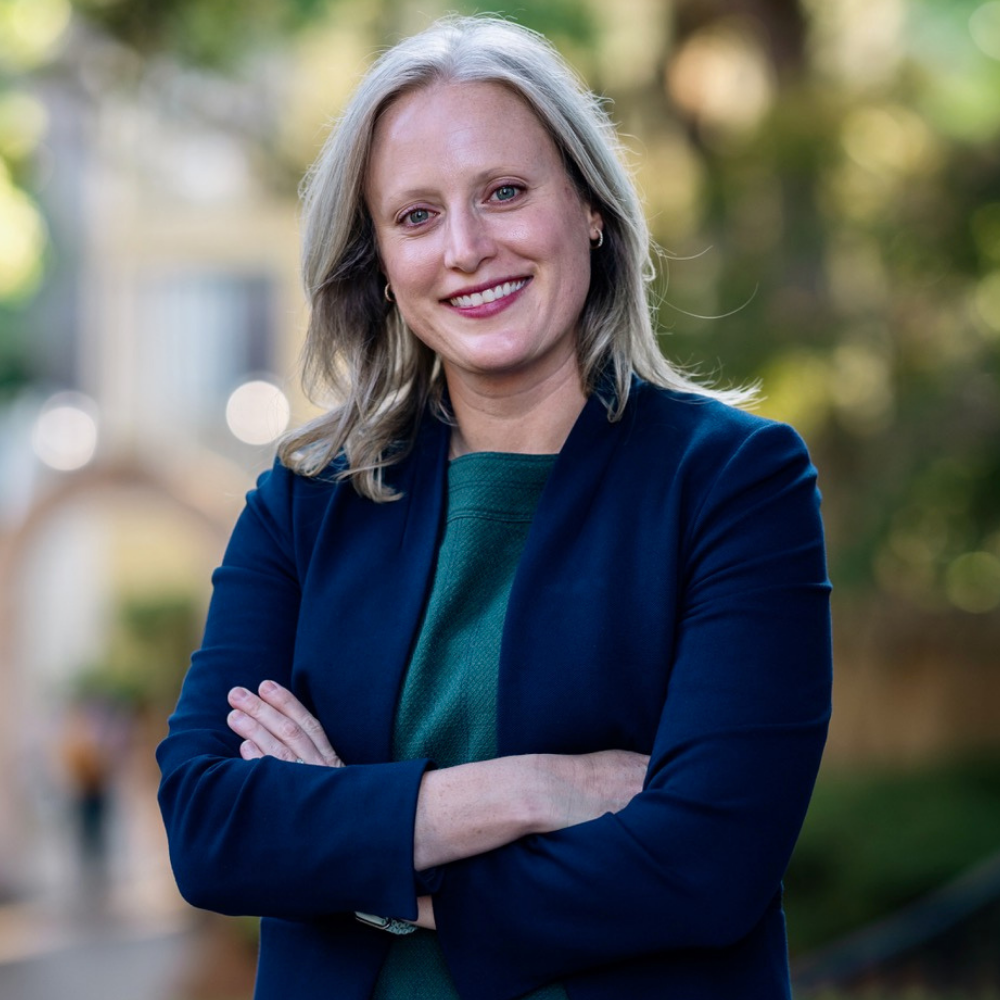 Professional outdoor headshot of a person smiling with arms crossed, dressed in a navy blazer and green top, with a softly blurred background