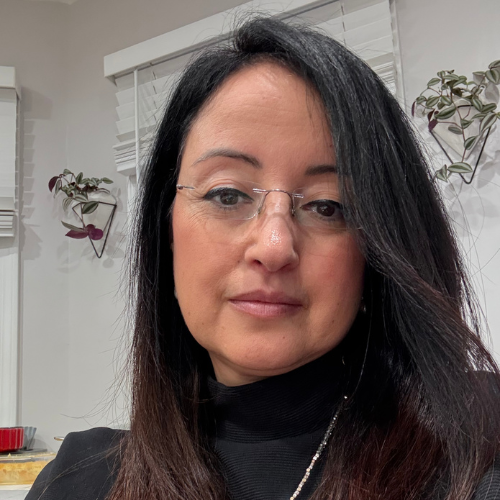 Close-up headshot of a person with long dark hair and glasses, wearing a black top, indoors with window blinds and small plants in the background.
