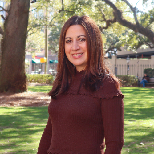 A woman with dark hair and dark sweater smiling outside on a sunny day.