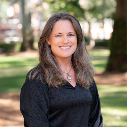 Wendy with long wavy hair wearing a black blouse and silver necklace, standing outdoors.