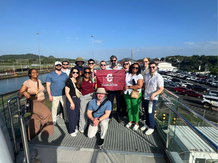 Group of adults standing on an elevated outdoor platform near a waterway and parking area, with one person holding a red banner that reads ‘Charleston.’ Clear blue sky and surrounding infrastructure visible in the background.