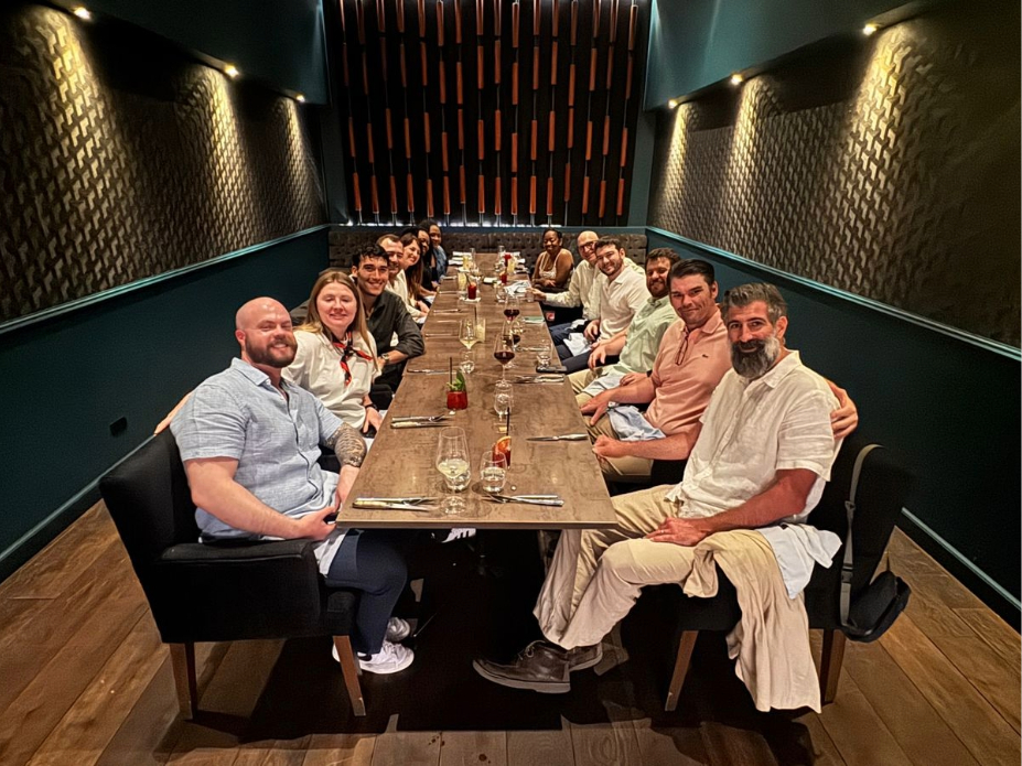 Group of adults seated along both sides of a long wooden table in a dimly lit private dining room, with place settings, glassware, and decorative wall panels visible.