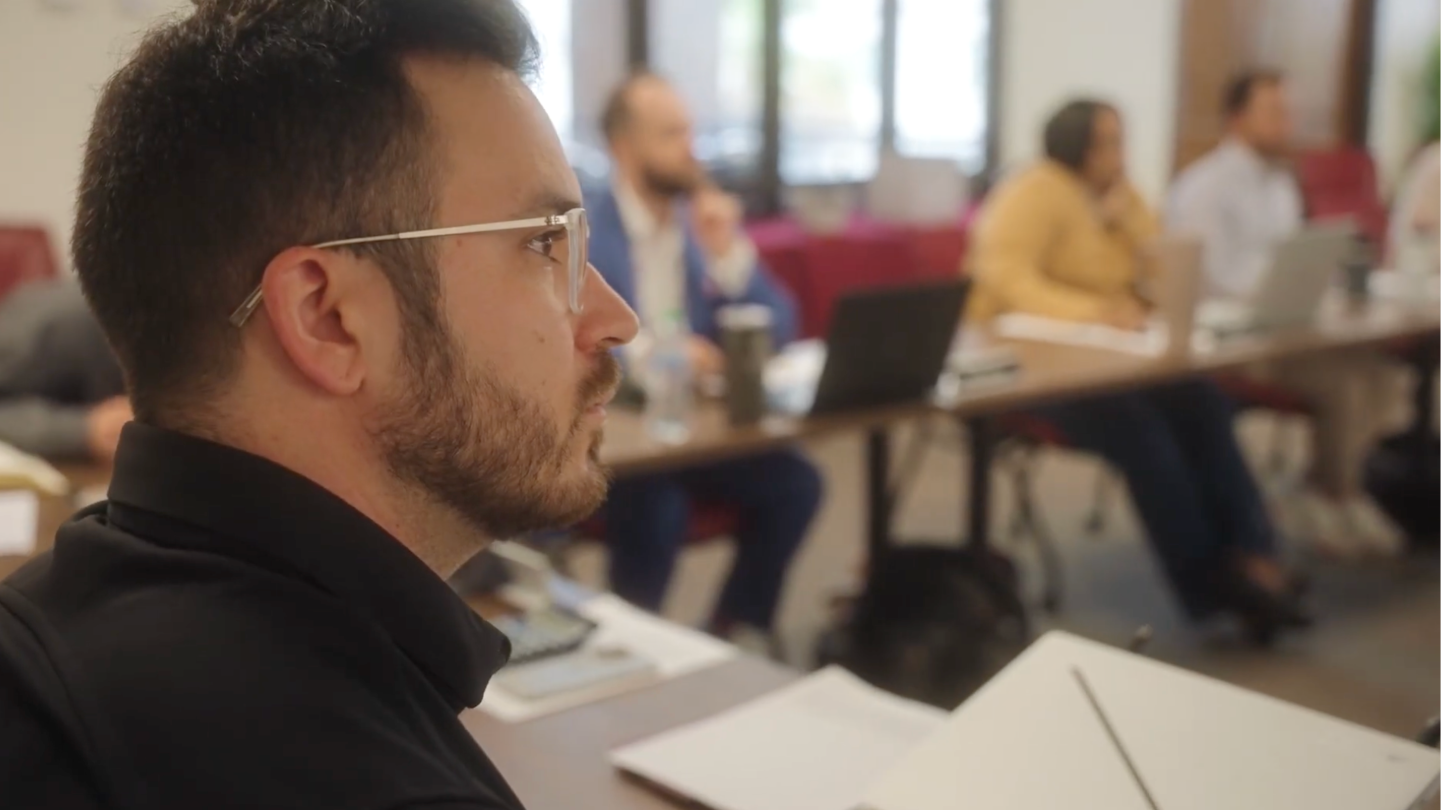 A closeup of a man with dark hair and glasses paying attention to someone speaking in a room