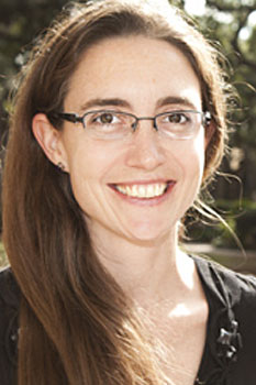 Professional headshot of a woman with long brown hair and glasses, smiling outdoors.