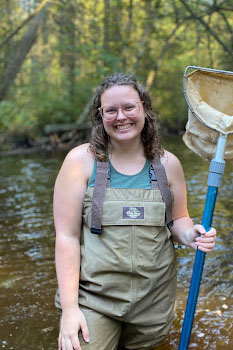 A person standing in a stream wearing waders and holding a net, surrounded by trees.