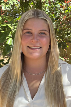A young woman with long blonde hair smiles warmly against a backdrop of greenery.