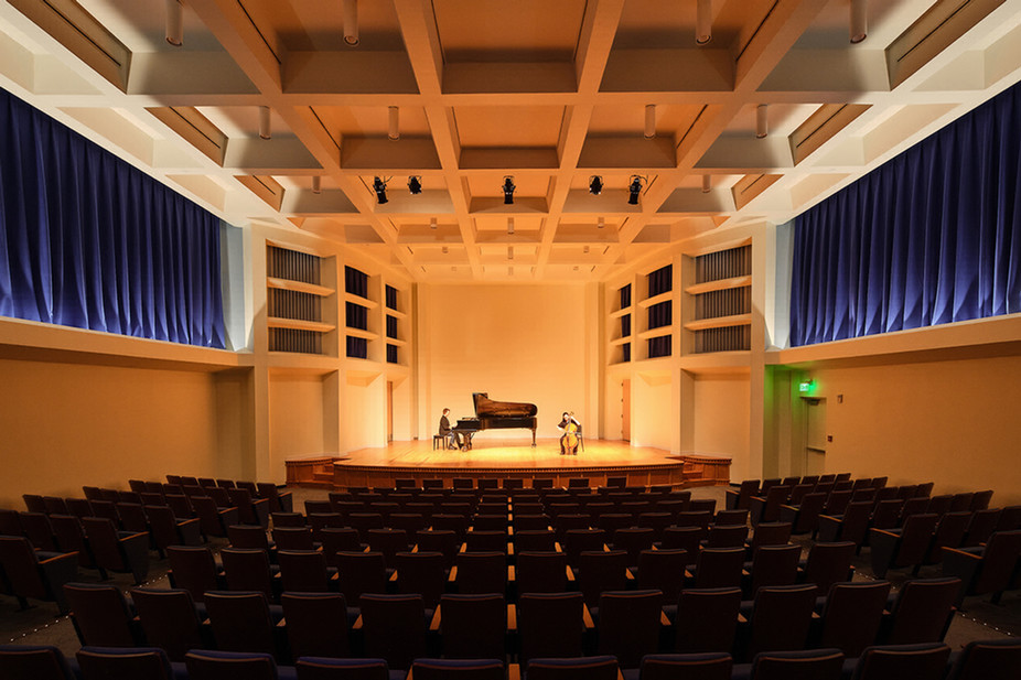 A wide view from the back of a concert hall of a pianist and cellist performing together on stage, with rows of seating in the foreground