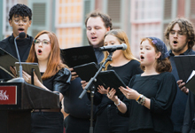 Choral group performing outdoors with sheet music, featuring diverse singers in black attire.