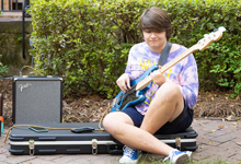 A young person playing an electric bass guitar while sitting on a guitar case outdoors.