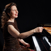 Angela Hewitt, smiling while seated at a grand piano, hands poised on the keys.