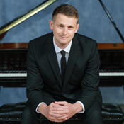 Tom Hicks, a man in a black suit sitting with hands clasped in front of a grand piano.
