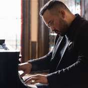 Jonathan Mamora in a dark jacket playing a piano in a softly lit room.