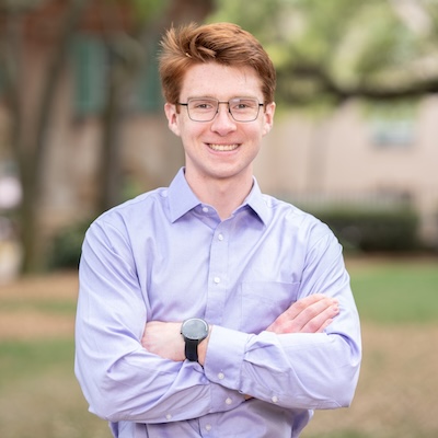 A smiling male student with glasses and a light blue button-down shirt, standing outdoors with arms crossed.