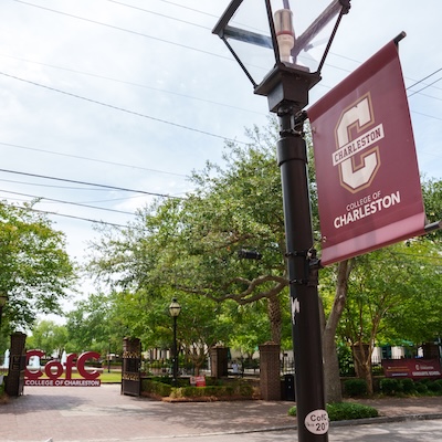 Campus entrance with a streetlamp banner reading “College of Charleston,” with trees and a gate in the background.