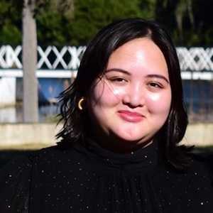 Woman smiling with shoulder-length dark hair wearing a black top outdoors, with a white fence and trees in the background.