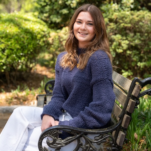Smiling young woman with long brown hair sitting on an ornate park bench, wearing a navy sweater and white pants, with greenery in the background.