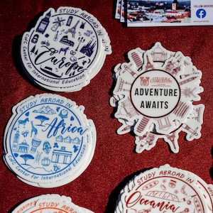 Travel-themed stickers on a red background, featuring a large “Adventure Awaits” badge surrounded by smaller “Study Abroad” stickers with illustrated landmarks.