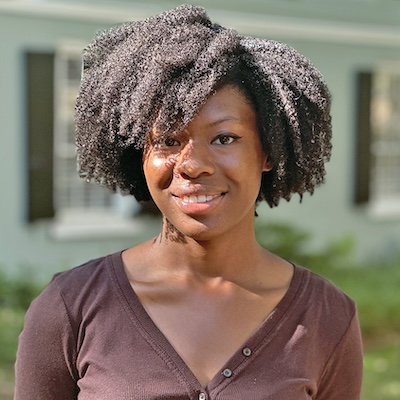 Smiling woman with natural curly hair standing outdoors in front of a house, with a softly blurred background.