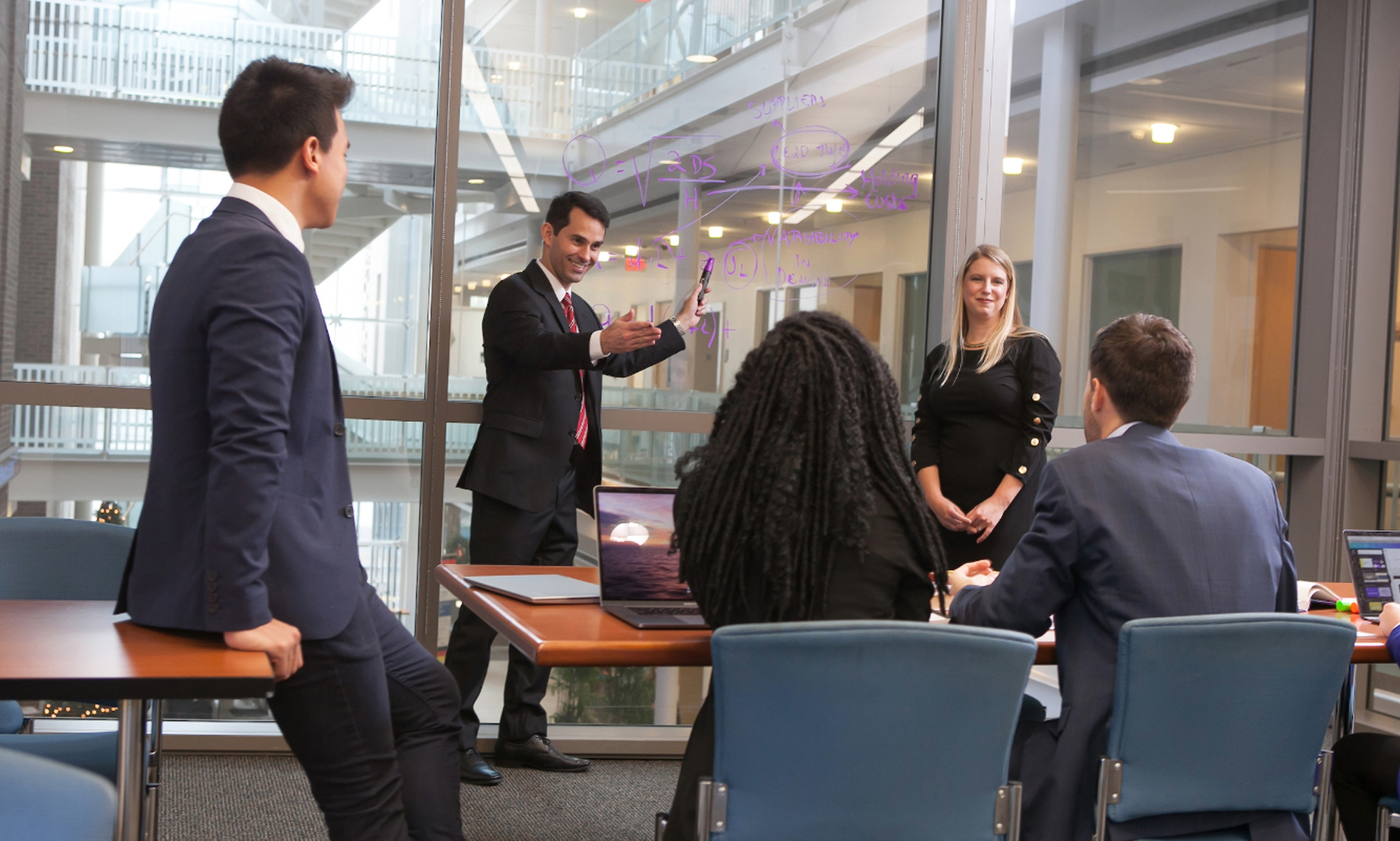 Business meeting in a glass-walled conference room as a presenter gestures toward notes written on the window while colleagues listen.