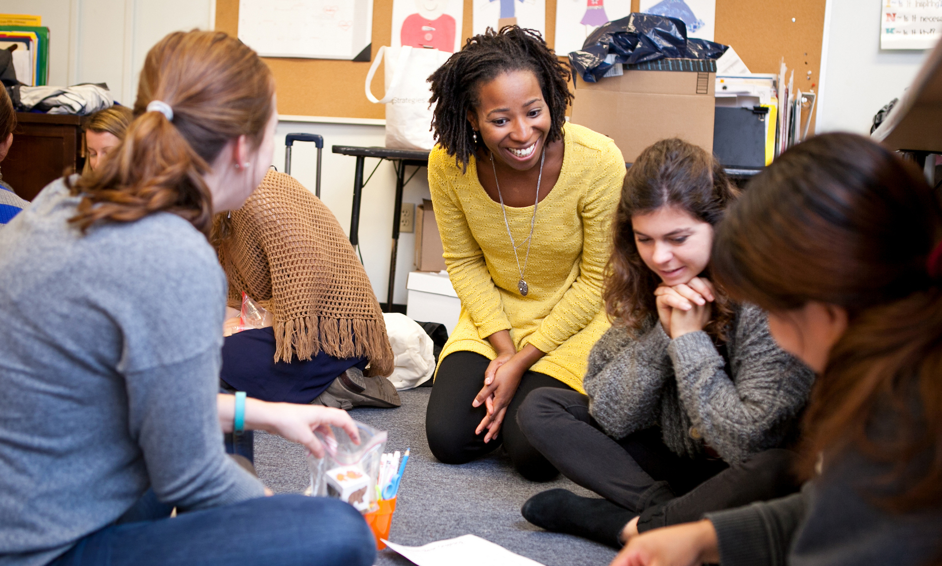 Teacher smiling and talking with a small group of students sitting on the classroom floor.