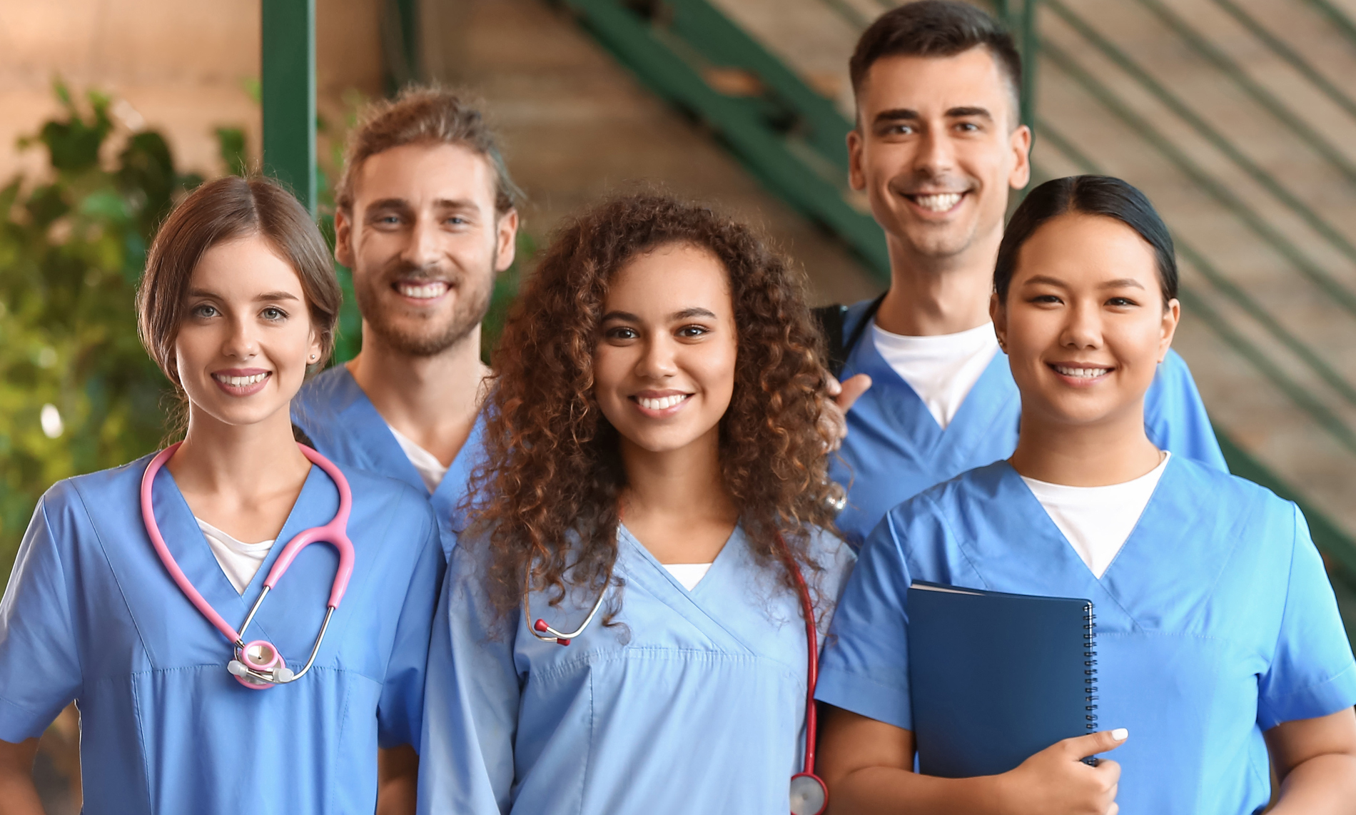 Smiling healthcare team in blue scrubs standing together, with stethoscopes and a notebook.