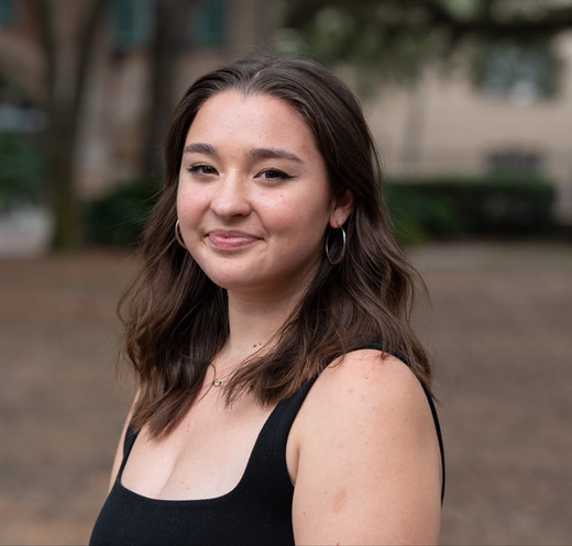 A smiling young woman with shoulder-length hair, wearing a black sleeveless top and hoop earrings, standing outdoors.