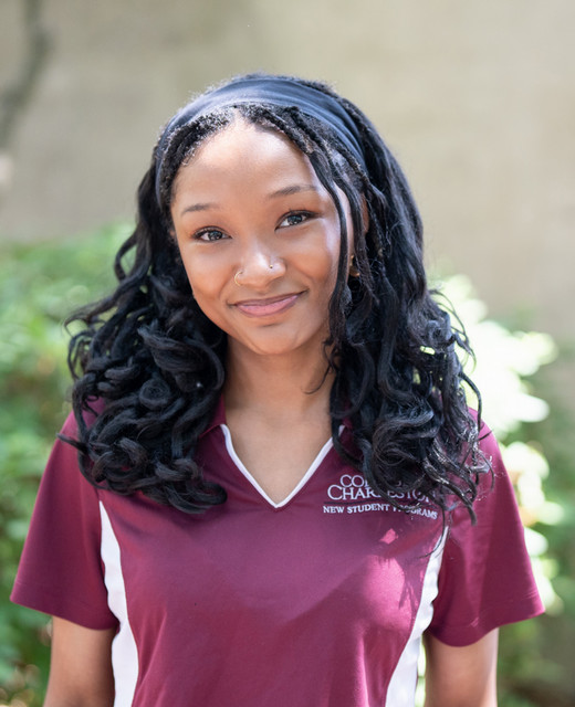 A young woman with curly hair wearing a maroon polo shirt, smiling confidently outdoors.