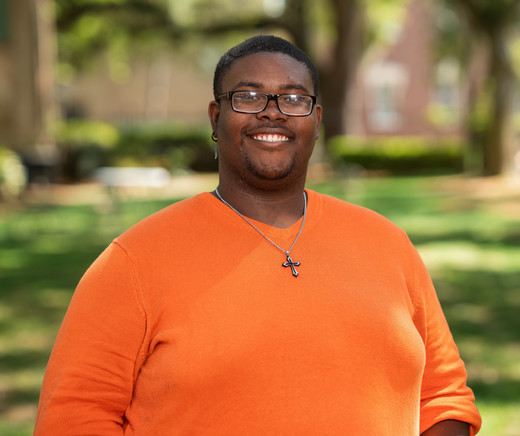 Smiling individual wearing an orange sweater, standing outdoors in a park-like setting with blurred background.