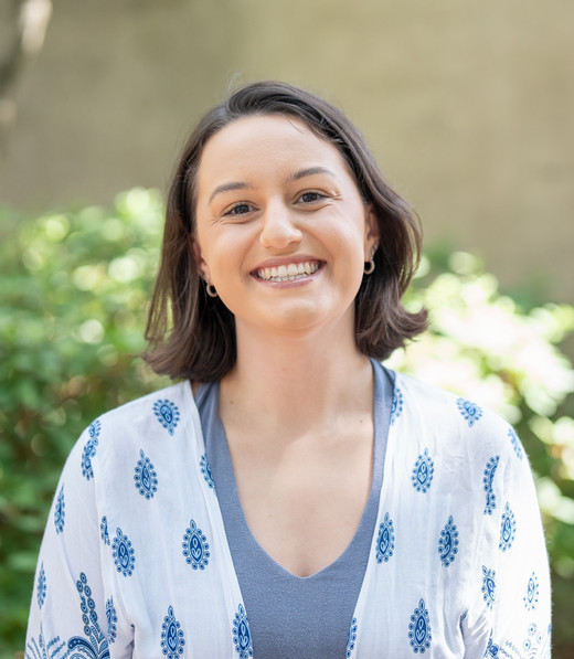 A smiling woman with shoulder-length brown hair is wearing a light blue top and a white patterned shawl, surrounded by greenery.