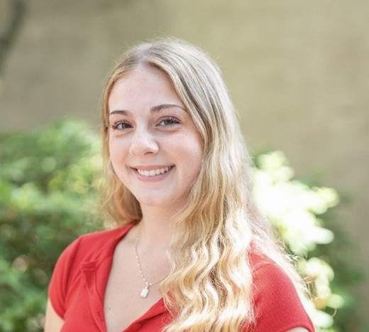 Young woman with long, wavy blonde hair wearing a red top, smiling in a natural outdoor setting.
